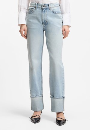 Jeans azzurri chiari a gamba dritta con ampi risvolti, indossati con scarpe slingback nere con punta affilata e camicia bianca a maniche lunghe.