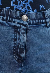 Vaqueros de denim azul con un botón decorativo plateado, lavado oscuro con una textura sutil y costuras estándar a lo largo de la cintura y los bolsillos.