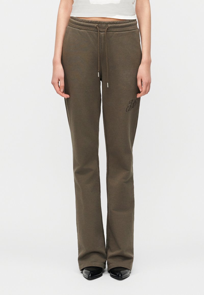 Καφέ ίσια φόρμα με κορδόνι στη μέση και διακριτι κό κεντητό γράμμα στο πάνω μέρος του μηρού, φορεμένη με μαύρα μυτερά παπούτσια.