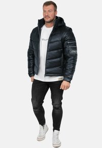 Giacca imbottita di un blu navy scuro con cappuccio, dotata di tasche con zip e polsini a costine. Indossata sopra una t-shirt bianca e abbinata a jeans neri e sneakers bianche.