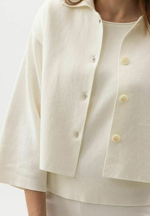 Nő krémszínű, gombos, rövid szabású kardigánt visel, alatta hozzáillő ujjatlan felsővel, amelynek kerek a nyakkivágása.