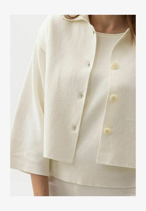 Kobieta ubrana w kremowy, zapinany na guziki krótki kardigan na ramiączkach, dopasowany do bezrękawnika z okrągłym dekoltem.