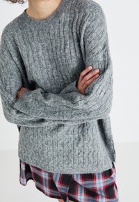 Person in einem strukturierten grauen Kabelstrickpullover und einem knielangen karierten Rock mit roten, schwarzen und blauen Mustern, Arme verschränkt.