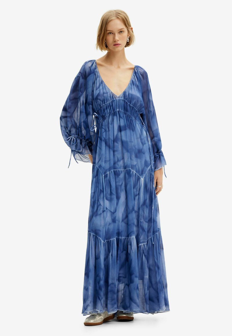 Maxikleid aus durchscheinendem blauen Stoff mit gebundenem Taillenbund, tiefem V-Ausschnitt, langen, luftigen Ärmeln und gestuften Rüschen am Saum. Silberne Schuhe.