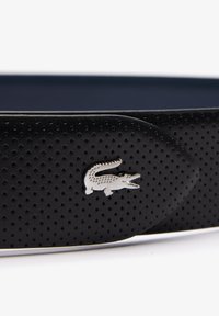 Cinturón de cuero negro con un diseño texturizado y perforado, acentuado con un logo de cocodrilo plateado. La hebilla es elegante y minimalista.