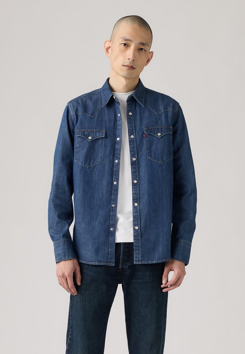 Camisa de mezclilla azul con botones a presión, que presenta dos bolsillos en el pecho y un diseño de yugo al estilo western. Llevada sobre una camiseta blanca.