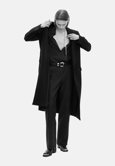 Eine Person in einem schwarzen Anzug mit weit geschnittenen Hosen, einem langen Mantel, schwarzen Stiefeln und geschichteten Perlenketten, die den Kragen des Mantels anpasst.