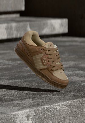Scarpa da ginnastica marrone e beige con lacci che fluttua sopra una superficie di cemento grigio e gradini sullo sfondo.