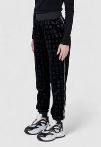 Pantaloni jogger in velluto nero con motivo a lettere ripetute, vita elastica e strisce laterali bianche, abbinati a sneakers chunky nere e bianche.