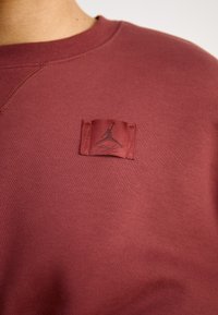 Bordeaux színű pulóver, melyen egy fekete logós szőtt címke található. Puha anyagú, texturált felületű és kerek nyakú.