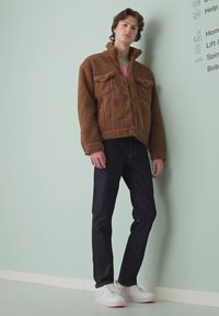 Bruine, fuzzy jas met twee borstzakken, zwarte jeans en witte sneakers met roze accenten, staand tegen een lichtgroene muur.