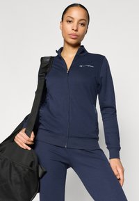 Ναυτικό μπλε αθλητικό μπουφάν με φερμουάρ, ψηλό γιακά και ριμπ μανσέτες, συνδυασμένο με αντίστοιχο παντελόνι· κρατάει μια μαύρη γυμναστηριακή τσάντα από δίχτυ.