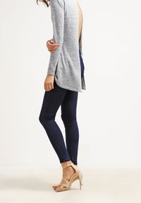 Cardigan gris à manches longues avec un dos ouvert, associé à un jean skinny foncé et des sandales à talons hauts beiges. Texture lisse et design moderne.