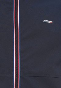 Μπλε μπουφάν με πλήρες φερμουάρ που έχει αντίθετη μαύρη ταινία και κόκκινες, λευκές και μπλε ρίγες, μαζί με ένα μικρό λογότυπο στο στήθος.