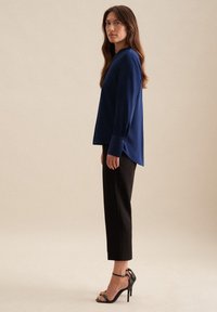 Blusa blu a maniche lunghe con collo alto e polsini abbottonati, abbinata a pantaloni neri a sigaretta e sandali neri con tacco alto.