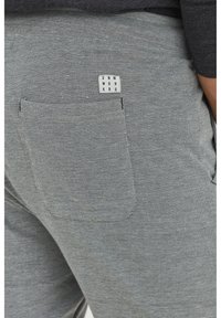 Pantalones grises con una tela texturizada, con un bolsillo trasero que muestra una etiqueta rectangular que dice "DNM WORKS".
