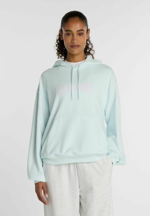 Frau mit lockigem Haar trägt einen hellblauen New Balance Hoodie und eine hellgraue Jogginghose und steht vor einem schlichten weißen Hintergrund.