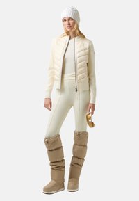 Pufferjack in crème met een getailleerd ontwerp, gecombineerd met crème leggings en beige gewatteerde boots. Accessoires omvatten een witte gebreide beanie en zonnebril.