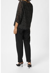 Blouse noire transparente à pois blancs, manches bouffantes trois-quarts, associée à un pantalon noir et des mules noires à talons.
