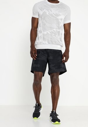 Homme portant une chemise blanche à manches courtes avec motifs, un short camouflage noir, et des chaussures de sport noires avec des rayures blanches et des semelles jaune fluo.