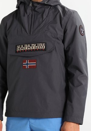 Ciemnoszara kurtka z kapturem z naszywką z logo Napapijri Geographic i emblem flagi Norwegii na piersi, noszona z jasnoniebieskimi spodniami.