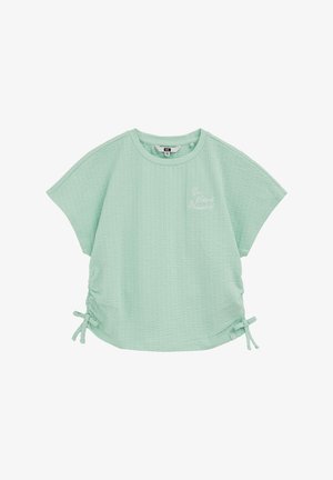 Haut texturé vert menthe à manches courtes avec col rond, liens à nouer sur les côtés et petit texte en script blanc sur le côté gauche de la poitrine.