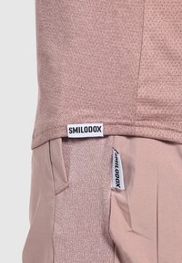 Nahaufnahme eines rosafarbenen, strukturierten Hemdes und einer Hose mit sichtbaren Smilodox-Markenetiketten an beiden Kleidungsstücken in der Nähe des Saums und der Tasche.