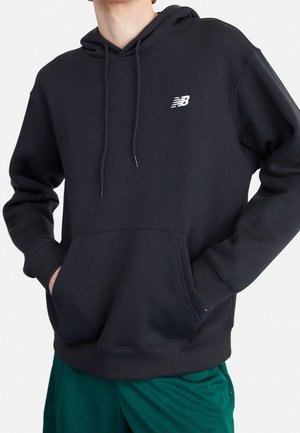 Osoba ubrana w czarną bluzę New Balance z kapturem, z kieszenią z przodu i sznurkami, w połączeniu z zielonymi sportowymi szortami, ręce w kieszeniach.