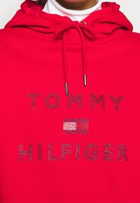 Felpa rossa con cappuccio regolabile e testo "TOMMY HILFIGER" in strass. Tessuto morbido con una vestibilità casual e dettaglio della bandiera del logo.