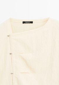 Haut en lin beige avec un décolleté en V, des fermetures latérales à boutons et une petite poche. Texture lisse, design minimaliste et esthétique décontractée.