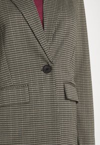 Hnědo-černý kostkovaný blazer, který má jeden černý knoflík, dvě přední kapsy a strukturovanou tkaninu. Klasické klopy.