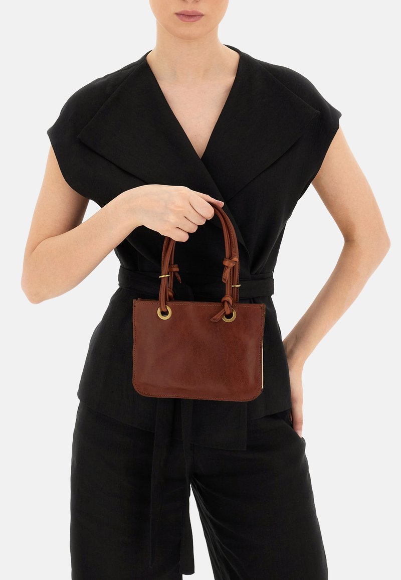 Borsa a mano in pelle marrone con due manici annodati, accenti in metallo dorato e forma rettangolare. Texture liscia, indossata da un modello in un outfit nero.