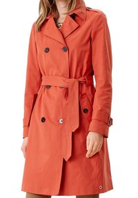 Oranžový trenčkot vyrobený z hladké tkaniny s dvouřadým zapínáním, černými knoflíky, páskem v pase a bočními kapsami.