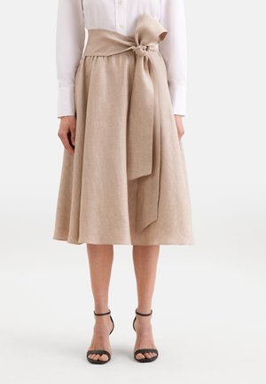 Beiger A-Linien-Midirock mit großer, vorne gebundener Schleife in der Taille, getragen mit einem weißen Langarmhemd und schwarzen Pumps mit Knöchelriemen.