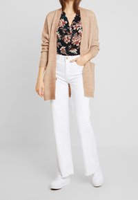 Cardigan in maglina beige, blusa nera floreale, jeans bianchi a gamba larga e sneakers bianchi. Il cardigan presenta tasche e una vestibilità rilassata.