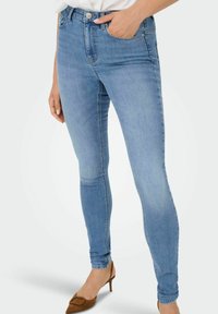 Sieviete, kas ģērbusies gaiši zilās šaurajās džinsās ar priekšējām kabatām, apvienojusi tās ar brūnām smailām kurpēm un baltu blūzi, roka kabatā.