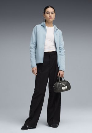 Femme portant un sweat à capuche zippé bleu clair, un haut blanc coupé, un pantalon large noir, des chaussures Puma noires, tenant un petit sac à main Puma noir.
