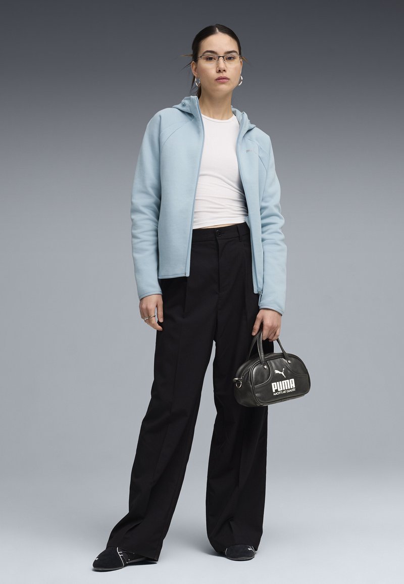 Femme portant un sweat à capuche zippé bleu clair, un haut blanc coupé, un pantalon large noir, des chaussures Puma noires, tenant un petit sac à main Puma noir.