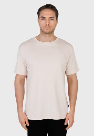 Mężczyzna z krótkimi brązowymi włosami, ubrany w prostą, jasnobeżową, krótką koszulkę i czarne spodnie, stojący na jednolitym białym tle.