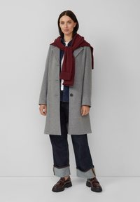 Grijze wolcoat met een opstaande kraag, bordeauxrode sjaal over de schouders, donkere spijkerbroek met opgerolde broekspijpen, en glanzende donkere schoenen.
