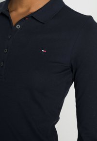 Marineblauw polo met lange mouwen, een gestructureerde afwerking, een knoopsluiting met drie knopen en een klein geborduurd logo in rood, wit en blauw op de borst.