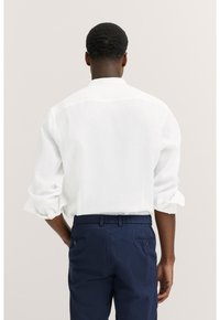 Weißes Leinenhemd mit hochgerollten Ärmeln, von hinten betrachtet. Getragen mit navyblauen Hosen mit elastischem Bund und rückseitigen Taschen.