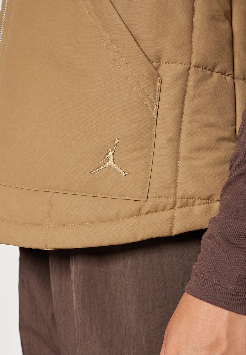 Beżowa kurtka puchowa z zamkiem z przodu, posiadająca boczne kieszenie i złote haftowane logo Jumpman na lewej części klatki piersiowej.