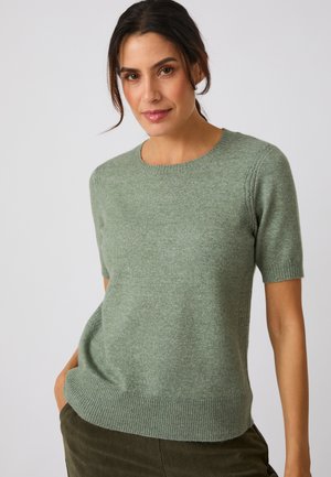Mujer con el cabello oscuro que lleva un suéter verde suave de manga corta y pantalones oscuros, de pie con una mano en el bolsillo sobre un fondo liso.