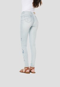 Gaiši zili plāni džinsi ar izšūtu ziedu dizainu uz kreisās kājas, klasiskā piecu kabatu stilā ar logotipa plāksnīti aizmugurē.