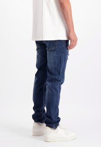 Een man in donkerblauwe jeans en witte sneakers, staande tegen een effen witte achtergrond, met gedeeltelijk zichtbare romp.