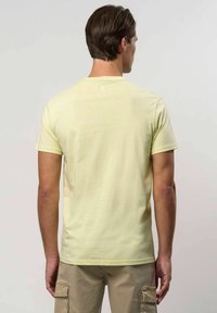 Rövid barna hajú férfi világos sárga, rövid ujjú pólót és bézs cargo sortot visel, háttal állva egy egyszínű háttér előtt.