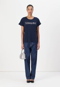 Mørkeblå T-skjorte med "Blauer" i metallisk skrift, kombinert med høy midje, løse blå jeans, holder en liten sølvveske, svarte sko.