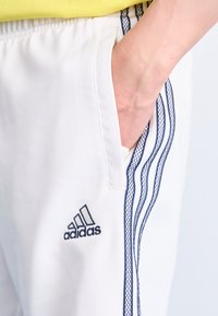 Bijele atletske hlače s mornarsko plavim trostrukim uzorkom, teksturiranim detaljima na džepovima i malim crnim Adidas logotipom s lijeve strane.