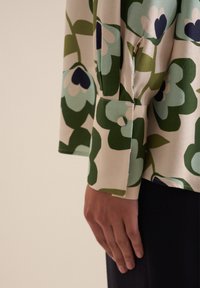 Blusa a fantasia floreale in crema con sfumature di verde e blu. Presenta una vestibilità ampia, polsini con bottone e una texture liscia.